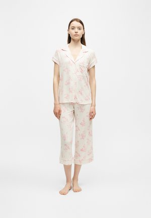 Jeune femme debout pieds nus portant un haut de pyjama blanc à manches courtes à boutons et un pantalon court à imprimé floral rose clair.