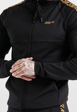Mężczyzna w czarnej bluzie z kapturem na zamek marki SikSilk z złotym logo i paskowymi detalami, ramiona skrzyżowane, pokazujące tatuaże na palcach i stawach palców.