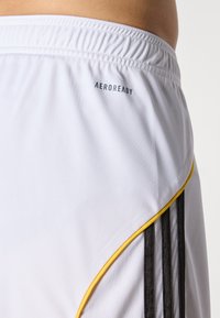 Shorts sportivi bianchi con vita elastica, logo "AEROREADY", righe verticali nere e piping giallo lungo la cucitura.