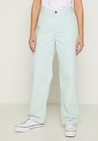 Jeans de perna larga em verde menta claro, feitos de denim, com fecho frontal, cinco bolsos e ténis brancos. Textura suave e ajuste descontraído.