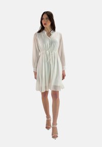 Vestido blanco de botones con cintura ajustable, mangas largas y transparentes, y falda fluida. Llevado con sandalias de tacón blancas.