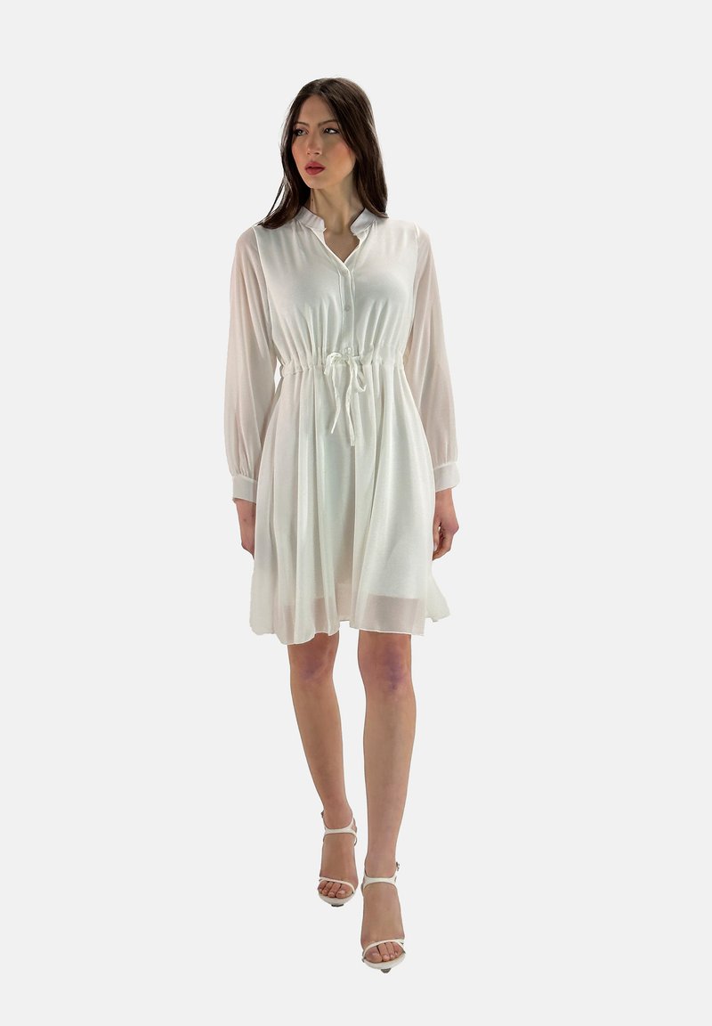 Vestido blanco de botones con cintura ajustable, mangas largas y transparentes, y falda fluida. Llevado con sandalias de tacón blancas.