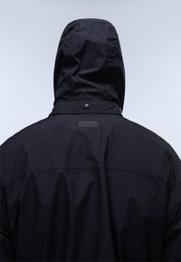 Person wearing a black hooded jacket viewed from behind against a plain light background.