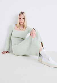 Glamžtais, ilgomis rankovėmis, mėtų žalios spalvos suknelė su kvadratiniu iškirpimu ir šonu plyšiu, derinama su baltomis slip-on bateliais ir glamžtomis kojinėmis. Auksinė grandinėlė.