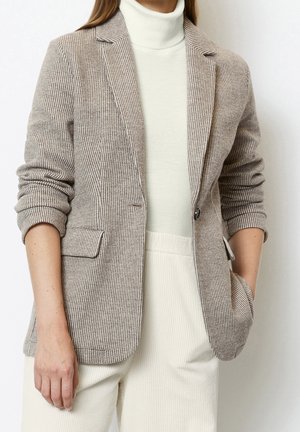 Vrouw draagt een beige pied-de-poule blazer over een witte coltrui en crème ribbroek, met één hand in de zak en opgestroopte mouwen.