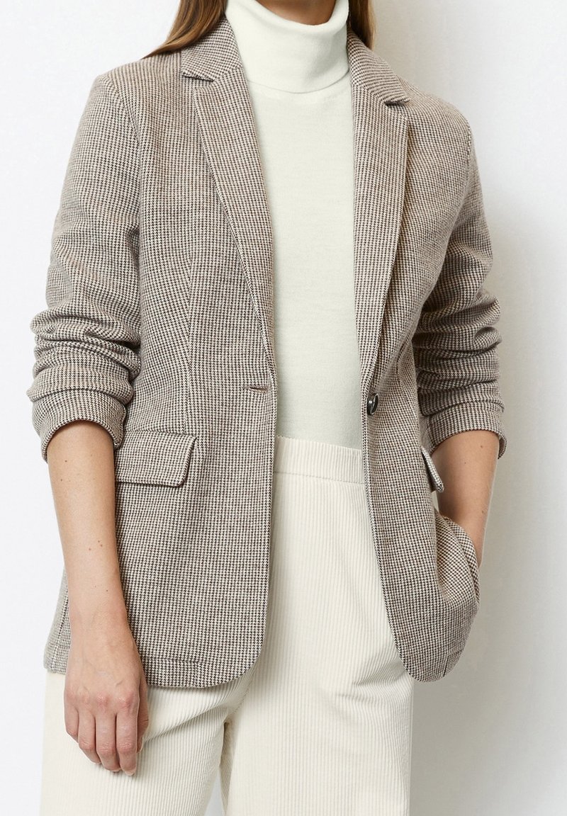Femme portant un blazer beige à motif pied-de-poule sur un col roulé blanc et un pantalon côtelé crème, une main dans la poche et les manches remontées.