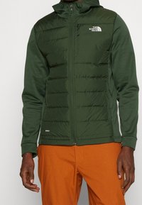 Vihreä puffatakki, jossa on sileä pinta, vetoketjullinen etuosa, huppu ja логотипin yksityiskohta. Takissa on vuoratut osat ja vartalonmyötäinen leikkaus.