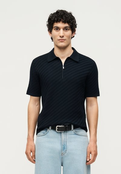 Homme aux cheveux bouclés portant un polo noir texturé à manches courtes et un jean bleu clair avec une ceinture noire, debout devant un fond uni.