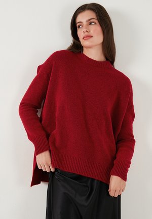 Vrouw in een losse rode gebreide trui en een zwarte rok, staand tegen een witte muur, kijkend naar de zijkant met haar handen bij de zoom van de rok.