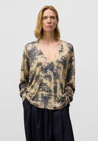 Beige langwerpige trui met lange mouwen en een V-hals, voorzien van marineblauwe stippenpatronen. Zachte textuur met gathere manchet en een relaxte pasvorm.