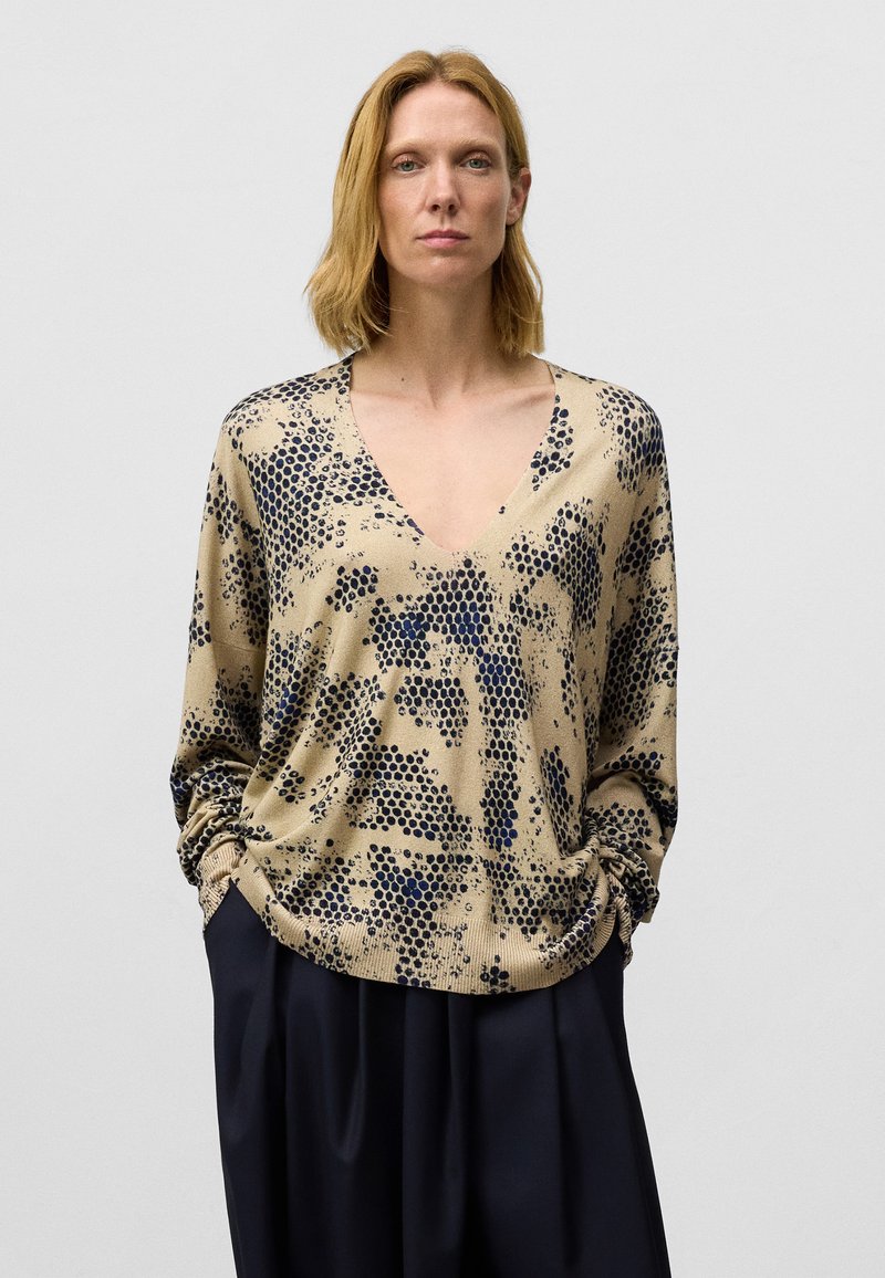 Beige langwerpige trui met lange mouwen en een V-hals, voorzien van marineblauwe stippenpatronen. Zachte textuur met gathere manchet en een relaxte pasvorm.