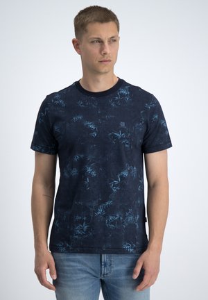 Młody mężczyzna w ciemnoniebieskim T-shircie z krótkim rękawem, z subtelnym jasnoniebieskim wzorem liści i jasnoniebieskich dżinsach, stojący na jednolitym tle.