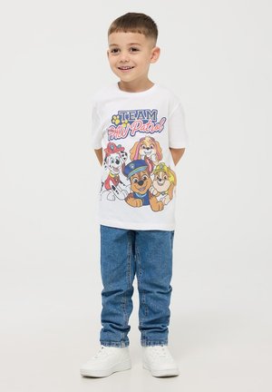 Maglietta bianca in cotone con cani dei cartoni animati colorati e la scritta "TEAM PAW Patrol", abbinata a jeans blu in denim e scarpe da ginnastica bianche.