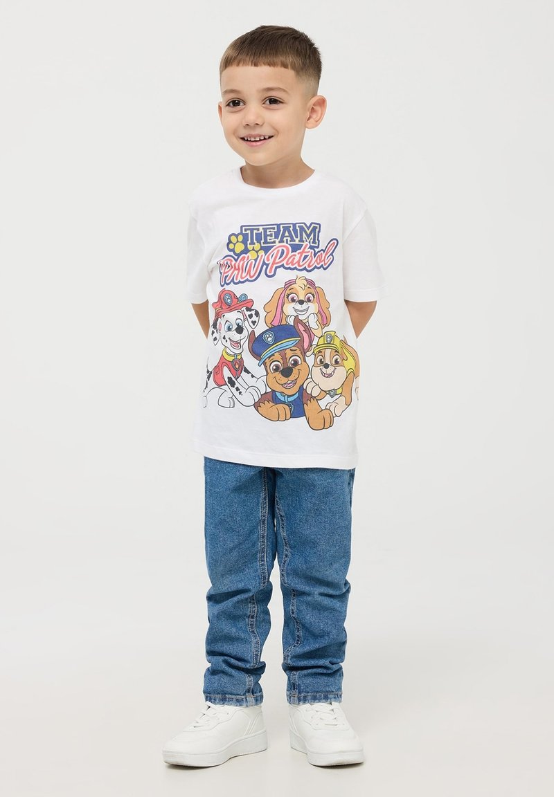 Maglietta bianca in cotone con cani dei cartoni animati colorati e la scritta "TEAM PAW Patrol", abbinata a jeans blu in denim e scarpe da ginnastica bianche.