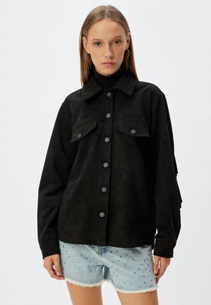 Schwarze Wildlederjacke mit Kragen, vorderem Knopfverschluss, zwei Brusttaschen und entspannter Passform; getragen über einem schwarzen Rollkragenpullover.