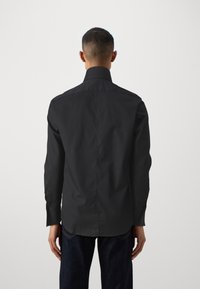 Černá košile s dlouhým rukávem, strukturovaným límcem a hladkým povrchem. Přiléhavý střih a manžety na knoflíky. Nosená s tmavými džíny.