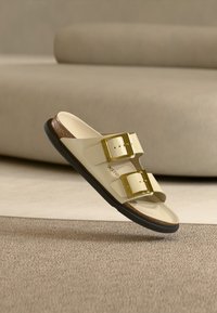 Ανοιχτό μπεζ σανδάλι slide με δύο αγκράφες σε χρυσή απόχρωση, υφασμένο καφέ πέλμα και μαύρη εξωτερική σόλα. Μινιμαλιστικός σχεδιασμός και ανοιχτή μύτη.