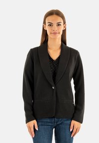 Zwarte blazer van textuurstof, met een sjawlcollar, twee voorzakken en een enkele knoopsluiting. Dragen over een top met stippen.