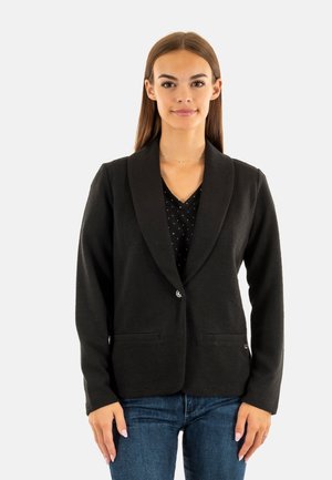 Blazer nero realizzato in tessuto testurizzato, con colletto a scialle, due tasche frontali e chiusura con un unico bottone. Indossato sopra una maglietta a pois.