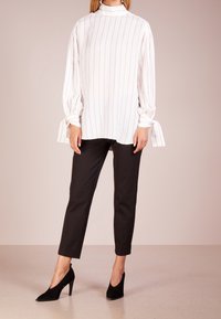Femme portant un haut blanc à col montant avec des rayures rouges et des poignets noués, un pantalon noir slim et des chaussures à talons hauts noires, se tenant sur un sol beige.