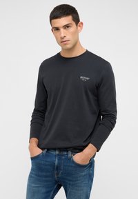 Juoda ilgomis rankovėmis marškinėliai su apvalia apačia ir balta prekės ženklo logotipu krūtinėje, derinami su mėlynais džinsais. Lygi tekstūra.