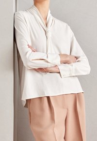 Bílá blůza s uzlíkem na límci, dlouhými rukávy a knoflíkovými manžetami. Slučuje se s měkkými, světle růžovými kalhotami.