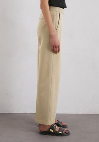 Pantaloni beige a gamba larga con una texture liscia, caratterizzati da pieghe frontali e tasche laterali. Abbinati a sandali neri con dettagli in hardware dorato.