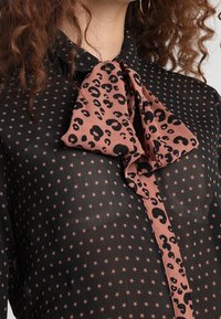 Schwarze Bluse mit Sternenmuster, ausgestattet mit einer großen, pinkfarbenen Leopardenmuster-Schleife am Kragen, die einen kontrastierenden Textur- und Designakzent setzt.