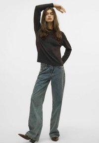 Maglione scuro con texture abbinato a jeans larghi di un blu sbiadito. Stivaletti con tacco marroni slanciano l'outfit. Il modello posa con un braccio alzato.