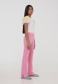 Top bianco con maniche a volant pastello, abbinato a pantaloni rosa a vita alta con spacchi laterali e un bottone dorato come dettaglio.