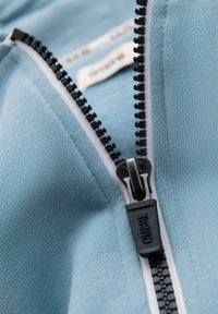 Gros plan sur une fermeture éclair noire sur un tissu bleu clair, partiellement fermée avec des dents visibles et une doublure en tissu blanc.