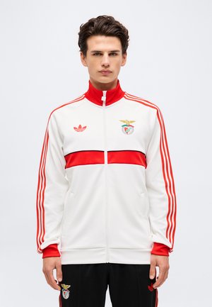 Ung mand iført en hvid og rød Adidas træningsjakke med Benfica-logo og matchende sorte bukser mod en ensfarvet baggrund.