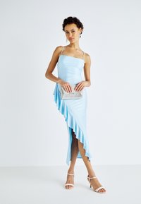 Robe ajustée bleu clair avec des fines bretelles, dotée d'un ourlet asymétrique froncé et d'une fente. Associée à une pochette à fermoir en argent et des talons légers.