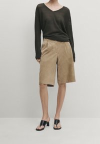 Ciemnozielony sweter w serek, o splocie dziewiarskim, zestawiony z jasnobrązowymi szortami ze zamszu, do kolan, z przednimi zakładkami, noszonymi z czarnymi sandałami na obcasie.