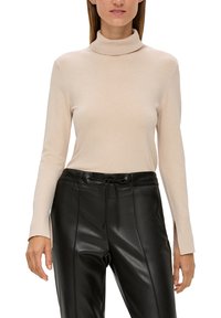 Beiger Rollkragenpullover mit langen Ärmeln, aus weichem Material, mit figurbetontem Design und seitlichen Schlitzen am Saum. Kombiniert mit schwarzen Lederhosen.