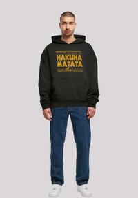 Felpa nera con testo "HAKUNA MATATA" in giallo, dotata di tasca frontale e polsini a costine, abbinata a jeans blu e sneakers bianche.