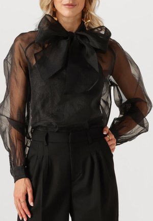 Zwarte doorschijnende blouse met een grote strik bij de kraag, doorschijnende luchtige mouwen en gecombineerd met hoge zwarte broeken. Glad van structuur.