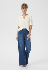 Witte top met korte mouwen en V-hals, gecombineerd met hooggeplaatste, wijde blauwe denim jeans. Schoenen zijn lichtbruine veterschoenen.