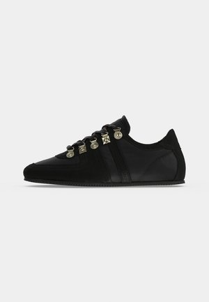 Zapatilla negra de corte bajo con paneles de ante y cuero, con ojetes decorativos metálicos y cordones negros sobre fondo blanco.