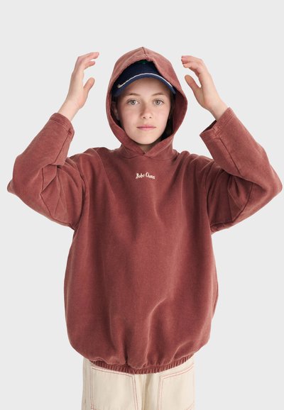 Junge Person trägt ein rostfarbenes Kapuzen-Sweatshirt über einer dunkelblauen Kappe und zieht die Kapuze mit beiden Händen zurecht, vor einem schlichten Hintergrund.