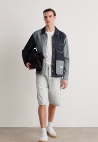 Jachetă din denim cu nasturi, în tonuri contrastante, pantaloni scurți albaștri deschiși și adidași albi. Țesătură texturată, buzunar cu clapă pe jachetă. Geantă neagră ținută în mână.