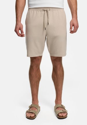 Mann trägt beige strukturierte Shorts mit Kordelzug und passende Wildledersandalen und steht vor einem weißen Hintergrund.