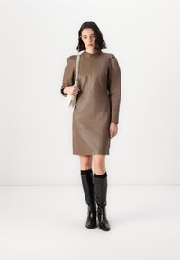 Rochie din piele maro cu umeri structurați, siluetă ajustată și lungime până la genunchi. Asortată cu cizme negre lucioase până la genunchi.
