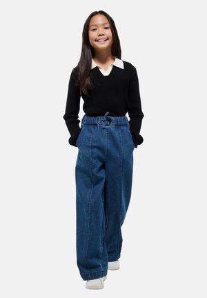 Jeune fille souriante, portant un pull noir avec un col blanc, un large pantalon en denim bleu et des baskets blanches, se tenant avec les mains dans les poches.