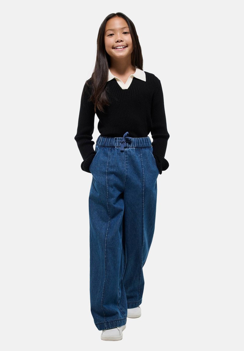 Jeune fille souriante, portant un pull noir avec un col blanc, un large pantalon en denim bleu et des baskets blanches, se tenant avec les mains dans les poches.