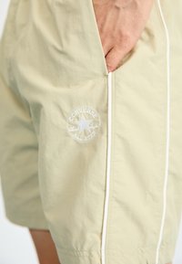 Lyse gule shorts laget av glatt stoff, med en hvit logo-lapp og sidesipperdetaljer. Enkel design med sømmer.