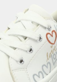 Λευκό αθλητικό παπούτσι με επάνω μέρος από λείο δέρμα, διακοσμημένο με γραφικά καρδιάς σε αντίθετο χρώμα και δίχρωμους κορδόνια. Ασημί μεταλλικά ανοίγματα για τα κορδόνια.