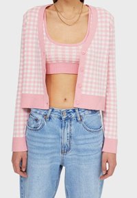 Růžový kostkovaný cropped cardigan s měkkou strukturou, s knoflíkovým zapínáním a ribbed manžetami, ležérně položený přes odpovídající bralette.