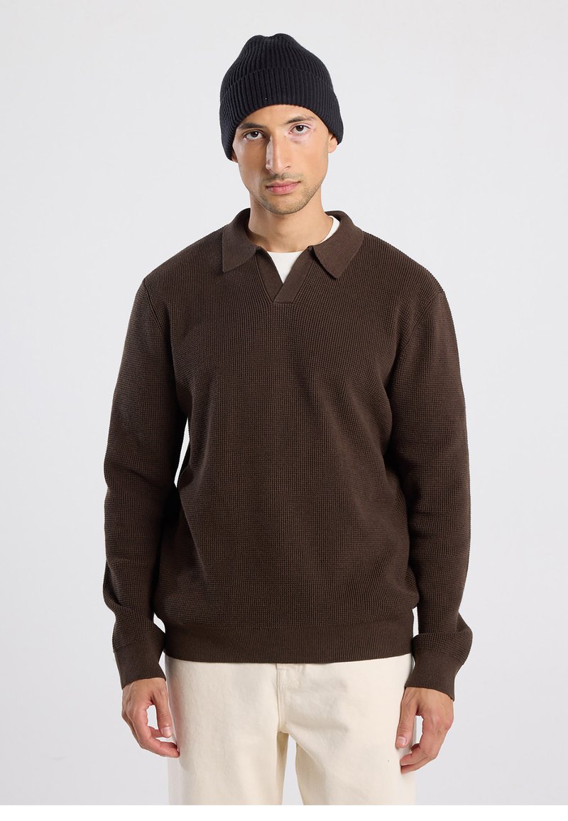 Pull marron tricoté avec col en V et col, associé à un bonnet côtelé noir. Le mannequin porte un pantalon de couleur claire, les bras détendus le long du corps.