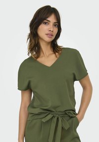 Blusa de manga corta color verde oliva con escote en V, fabricada en una suave textura y con un detalle de cintura atada. Ajuste casual, con un bolsillo lateral.